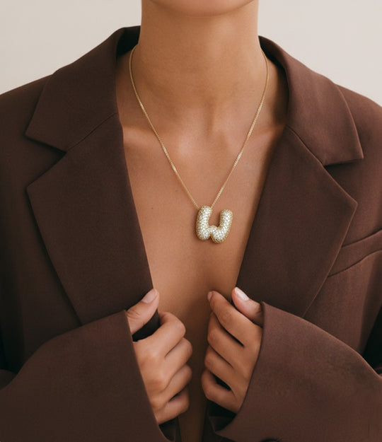 Close-up of a woman wearing a gold initial pendant necklace with a bubble letter design, styled with an open brown blazer.