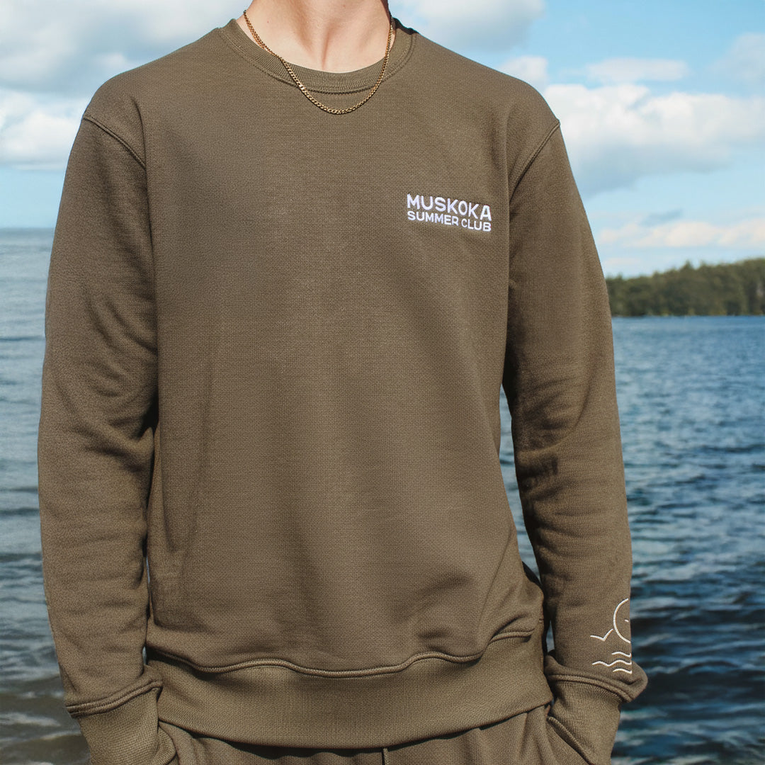 Person wearing a brown sweatshirt with 'Muskoka Summer Club' logo by a lake.