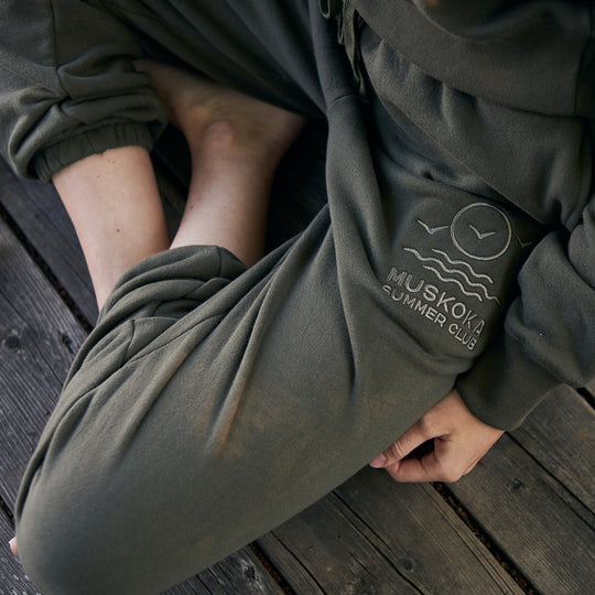 Person wearing green sweatpants with 'Muskoka Summer Club' logo on a wooden surface