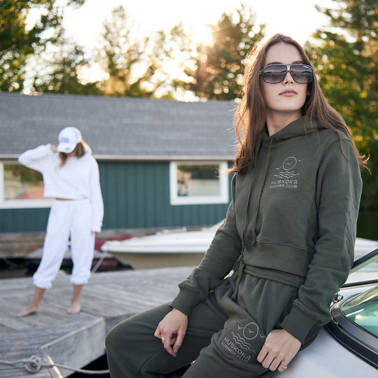 Woman wearing a green hoodie and pants sitting on a boat with another person in the background.
