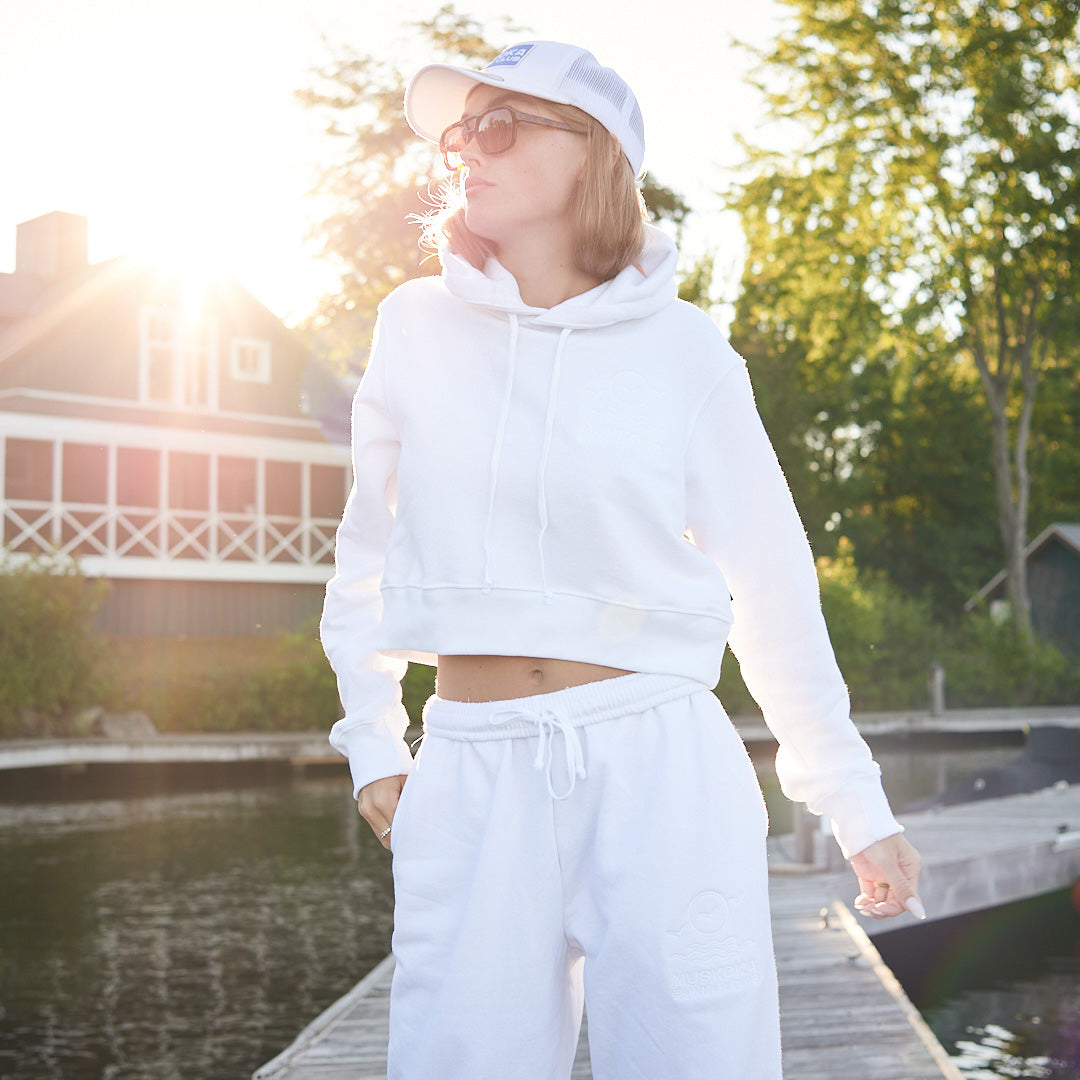Person wearing a white hoodie and pants standing on a dock with trees and a house in the background.