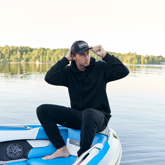 Man sitting on a boat wearing a black hoodie and cap with a lake and trees in the background