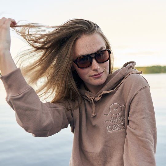 Woman wearing a brown hoodie with a logo, standing by the lake