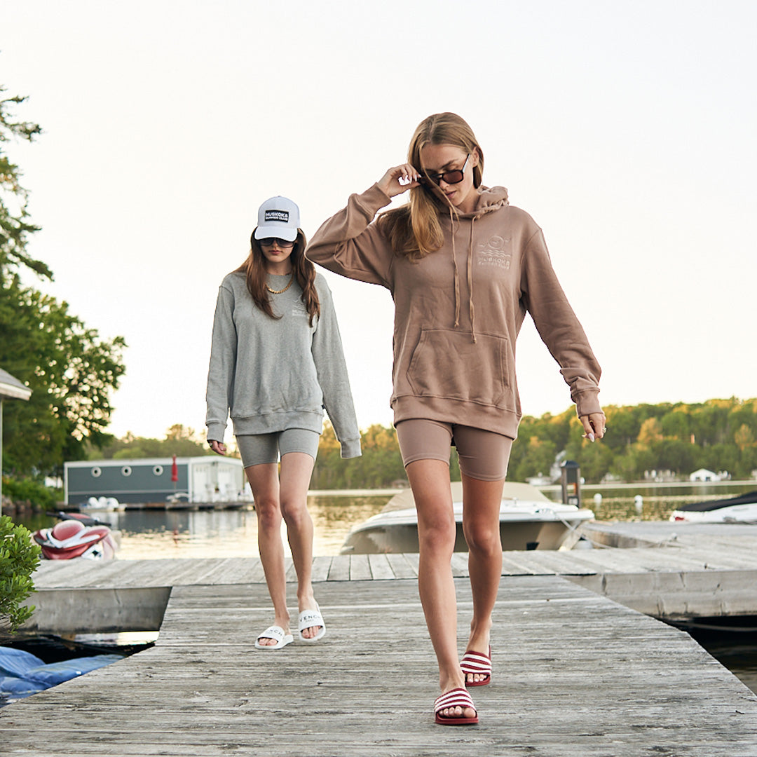 Two women walking on a dock by a lake, wearing hoodies and shorts.