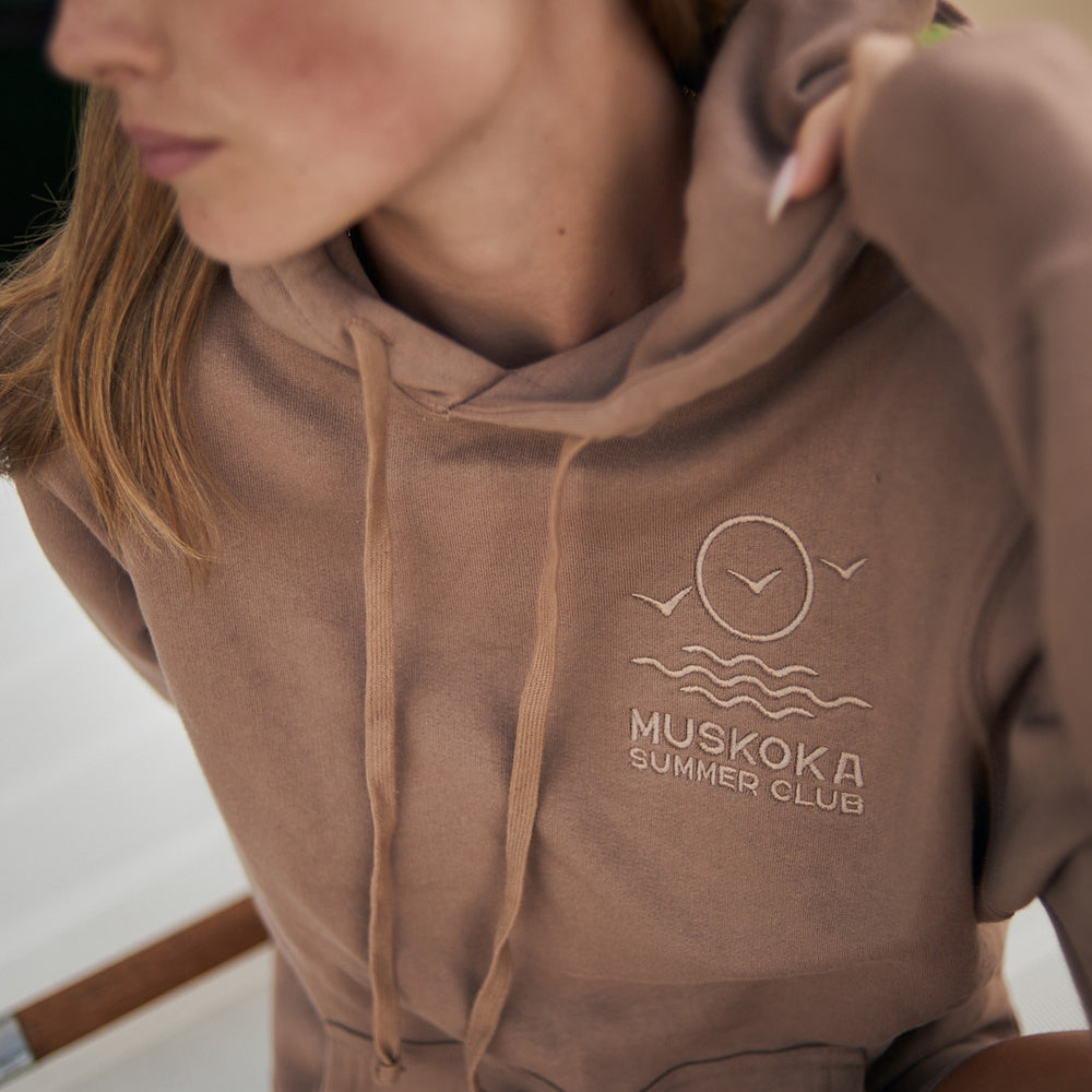 Brown hoodie with 'Muskoka Summer Club' logo worn by a person.