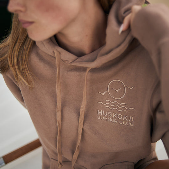 Brown hoodie with 'Muskoka Summer Club' logo worn by a person.
