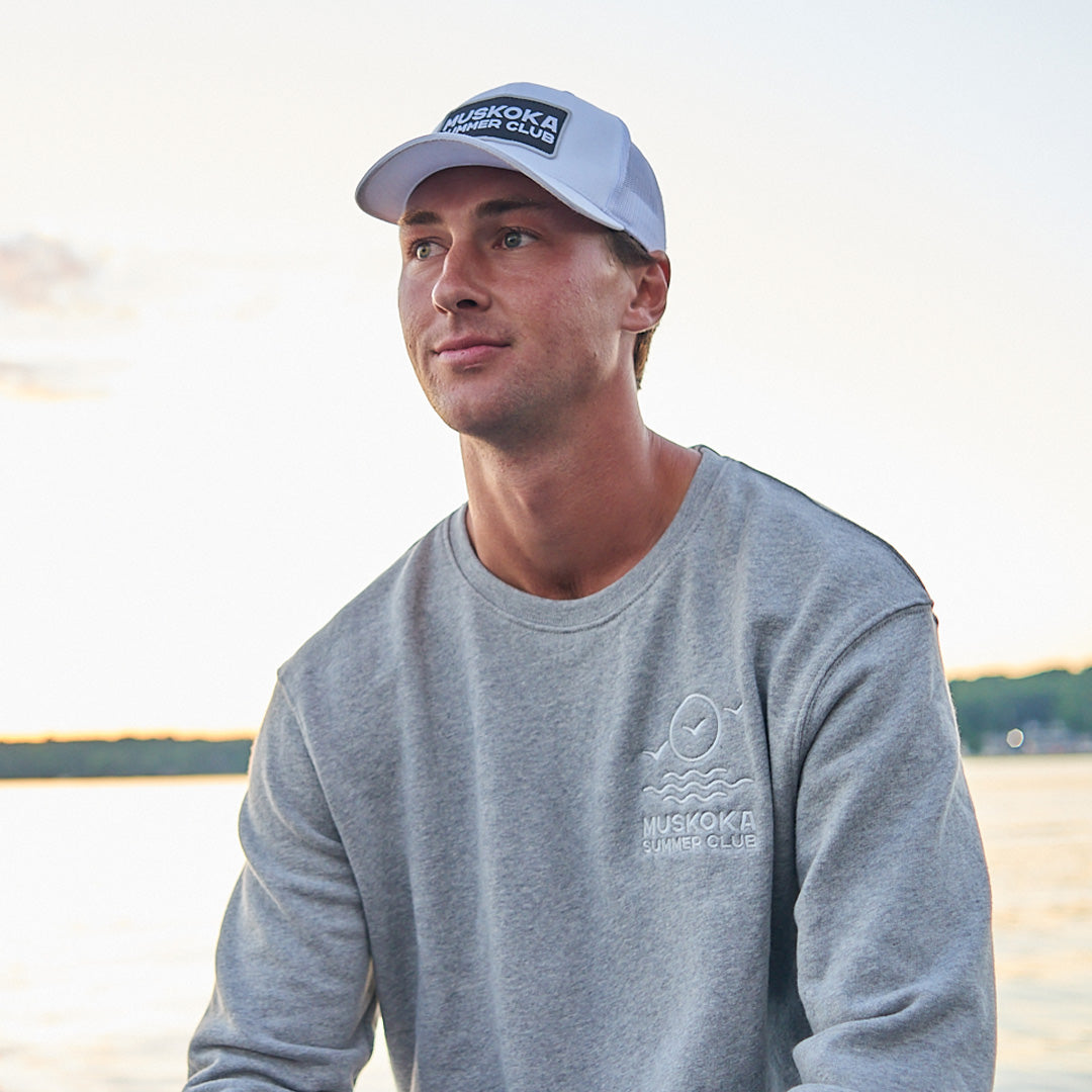 Man wearing a gray sweatshirt and cap with 'Muskoka Summer Club' branding by a lake.
