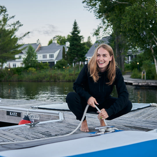 Person tying a rope to a boat on a dock with houses and trees in the background