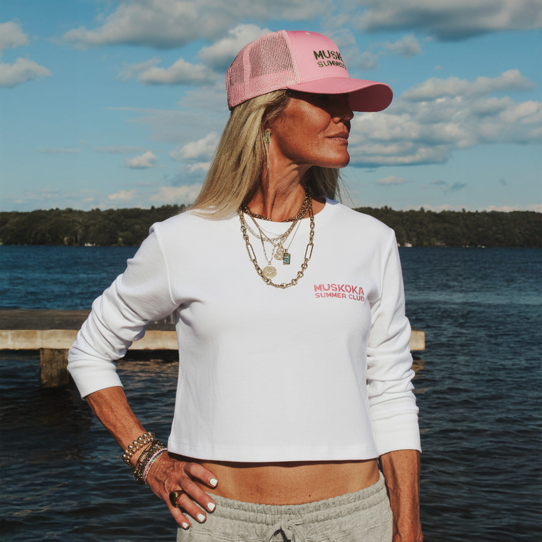 Woman wearing a white long-sleeve shirt and pink cap with Muskoka Summer Club branding by a lake.