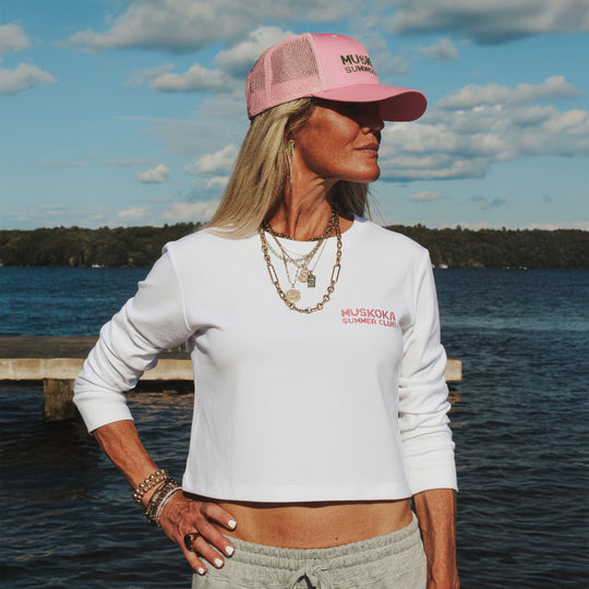 Woman wearing a white long-sleeve shirt and pink cap with Muskoka Summer Club branding by a lake.