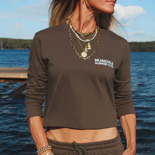 Person wearing a brown 'Muskoka Summer Club' longsleeve tee by a lake