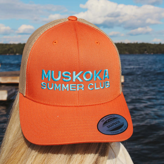 Orange cap with 'Muskoka Summer Club' text against a lakeside background