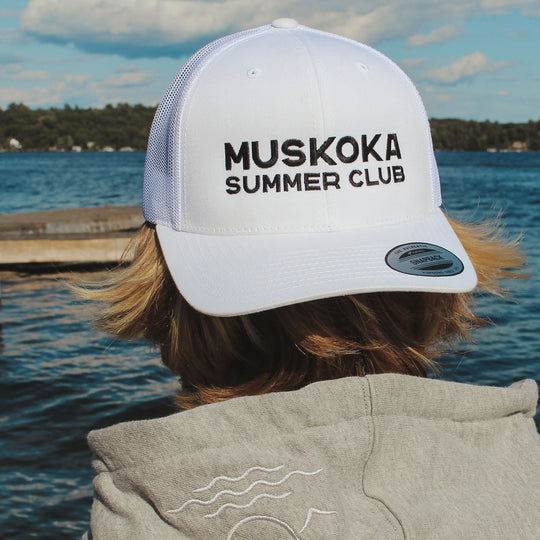 Person wearing a 'Muskoka Summer Club' cap by a lake