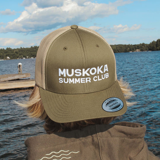 Person wearing a 'Muskoka Summer Club' cap by a lake
