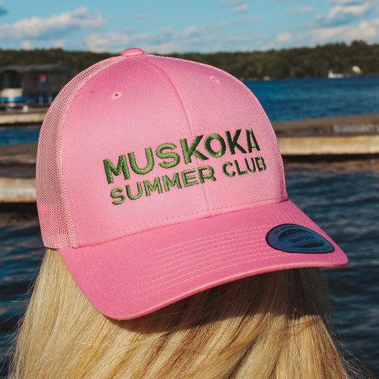 Pink cap with 'Muskoka Summer Club' text worn by a person by a lake.