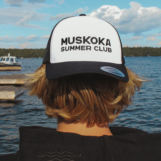 Person wearing a 'Muskoka Summer Club' cap by a lake with a boat and dock in the background.