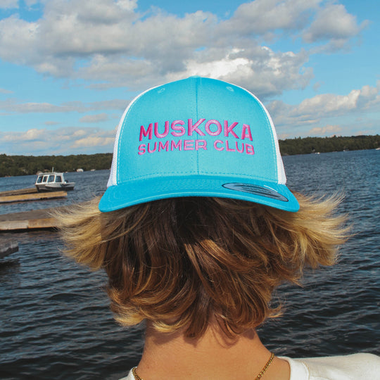 Person wearing a blue and white cap with 'Muskoka Summer Club' text, standing by a lake.