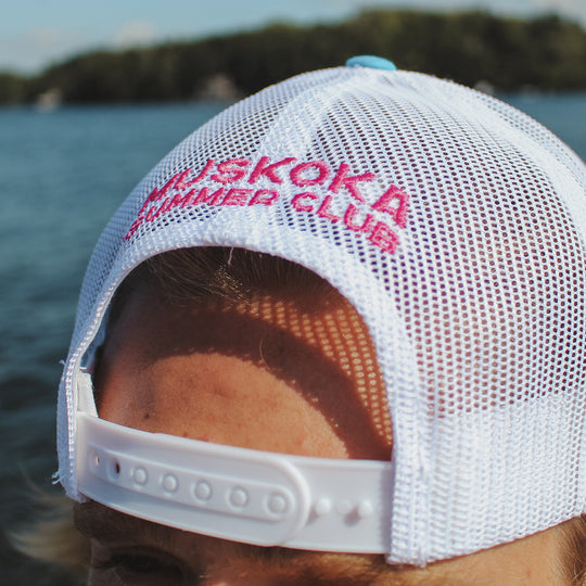 Person wearing a white mesh cap with ' Muskoka Summer Club' text against a water background