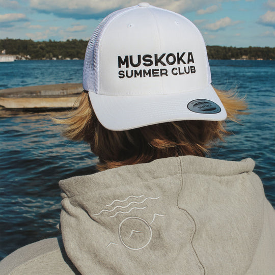 Person wearing a 'Muskoka Summer Club' cap and hoodie by a lake