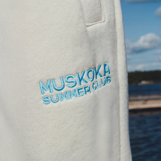 Close-up of a gray sweatshirt with 'Muskoka Summer Club' embroidery against a blurred natural background.