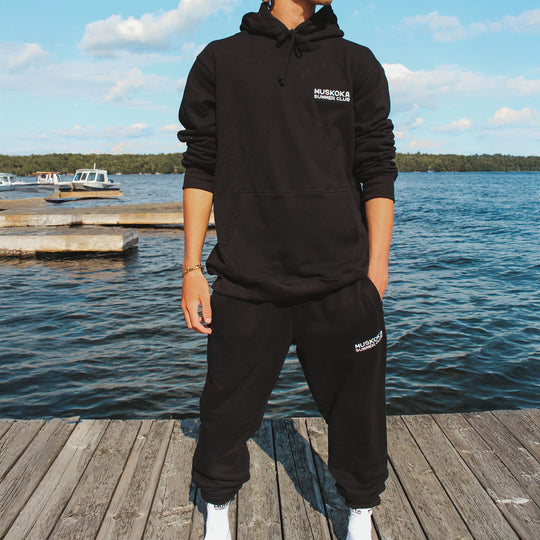 Person wearing a black hoodie and pants with a logo, standing on a dock by a lake.