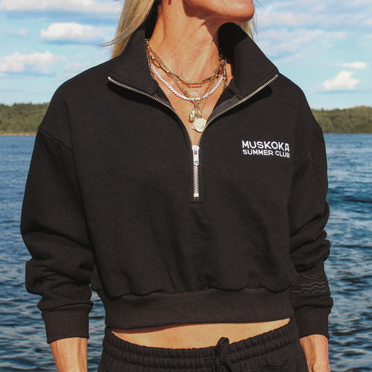 Person wearing a black 'Muskoka Summer Club' jacket by a body of water.