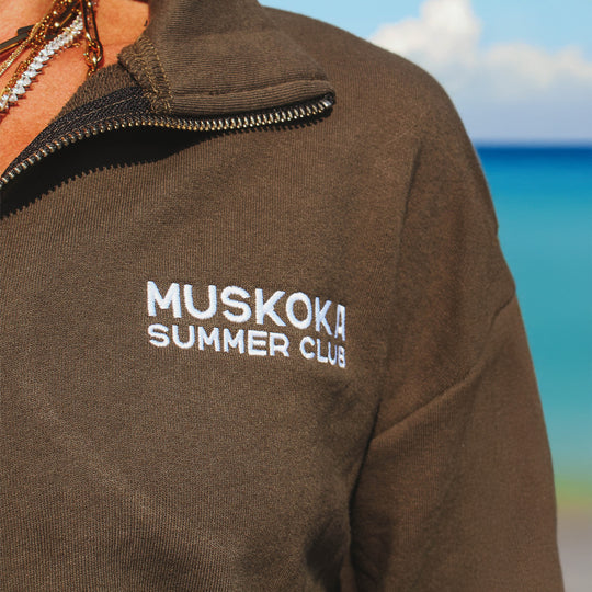 Brown jacket with 'Muskoka Summer Club' text on a blurred beach background
