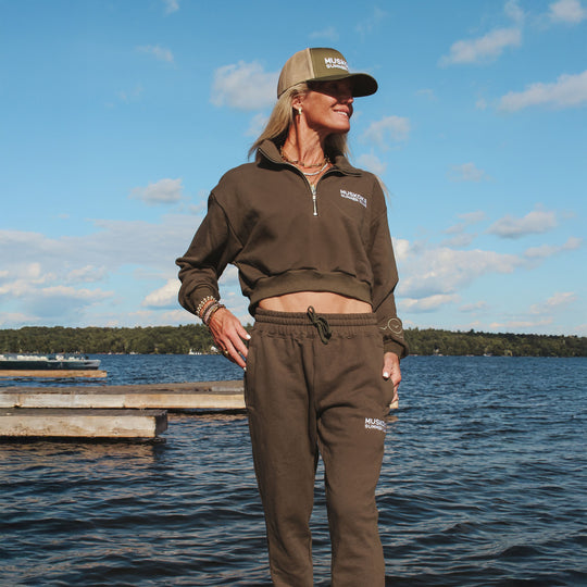 Person wearing a brown outfit with a cap standing by a lake