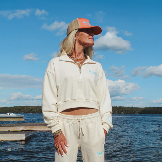 Woman in a beige sporty outfit and a cap standing by a lake with a clear sky.
