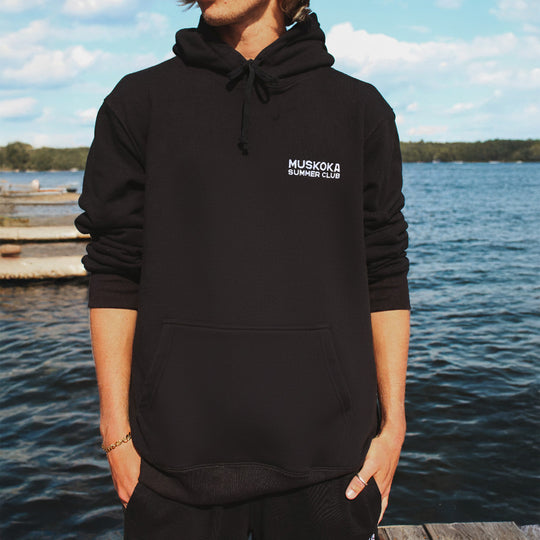 Person wearing a black hoodie with 'Muskoka Summer Club' logo by a lakeside.