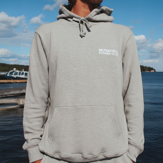 A person wearing a gray hoodie with 'Muskoka Summer Club' logo by a waterfront.