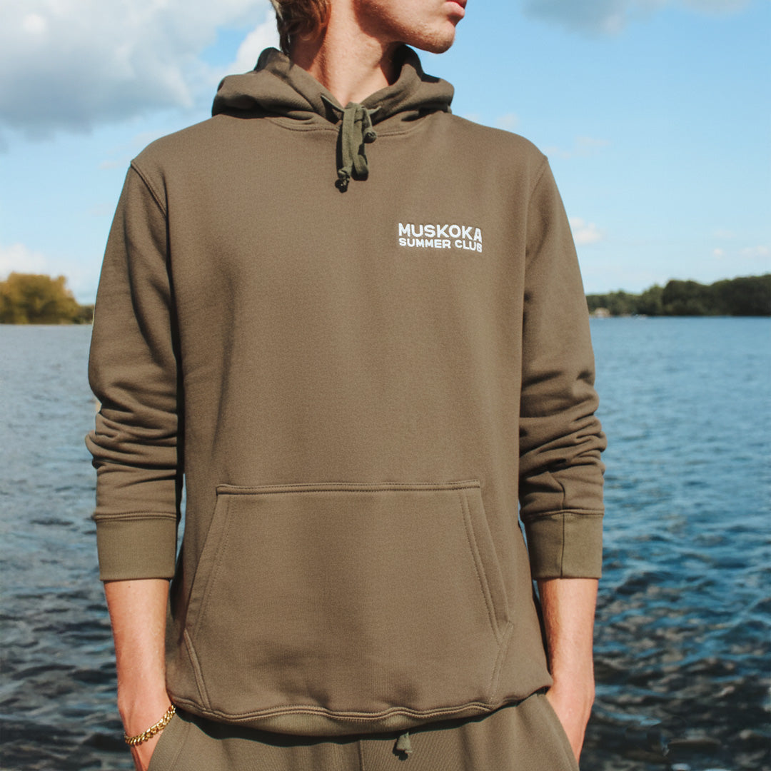 Person wearing a brown hoodie with 'Muskoka Summer Club' logo by a lake.