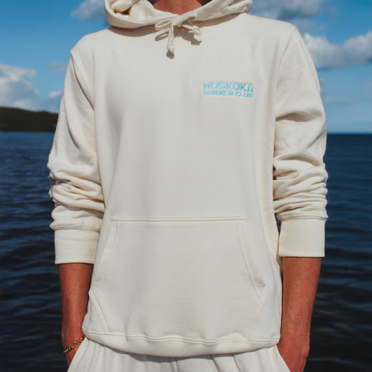 Person wearing a beige hoodie with a logo in front of a lake.