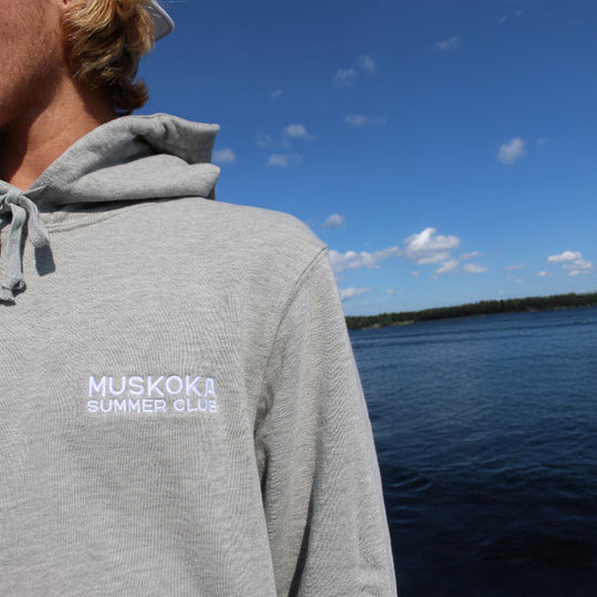 Person wearing a gray hoodie with 'Muskoka Summer Club' text, looking at a lake.