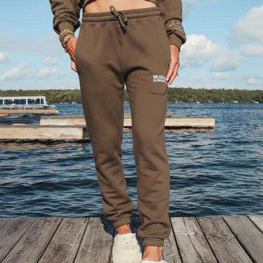 Person wearing brown sweatpants with a visible brand logo, standing on a dock by a lake.