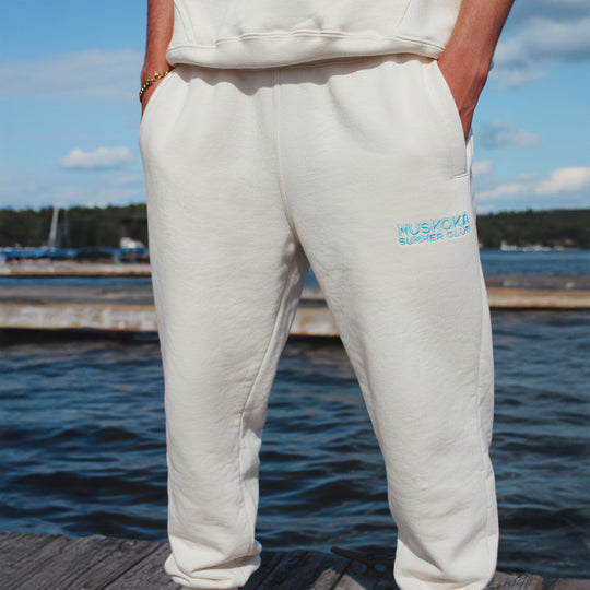 Person wearing white sweatpants with 'Muskoka Summer Club' logo by a lakeside.