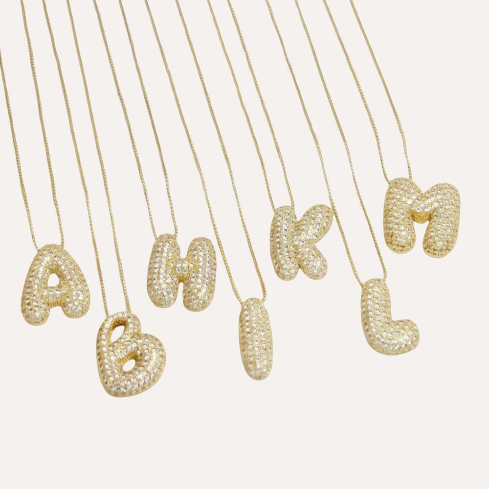 Gold bubble letter initial necklaces arranged on a white background, showcasing letters A through M with a sparkling diamond-like texture.