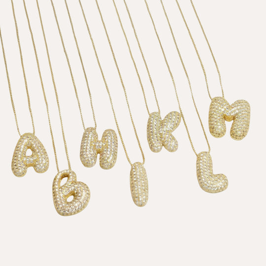Gold bubble letter initial necklaces arranged on a white background, showcasing letters A through M with a sparkling diamond-like texture.