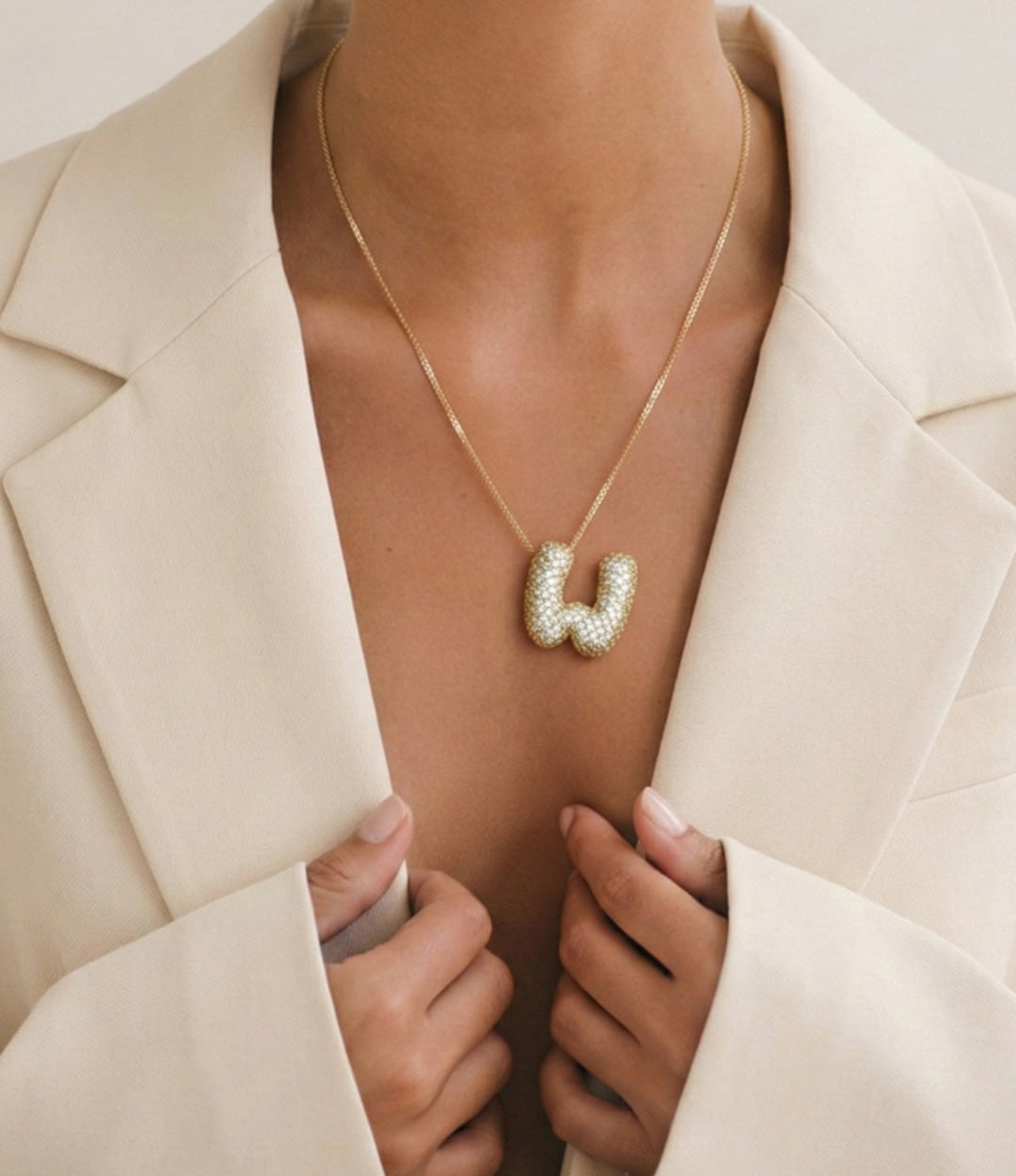 Close-up of a woman wearing a gold initial pendant necklace with a bubble letter design, styled with an open brown blazer.