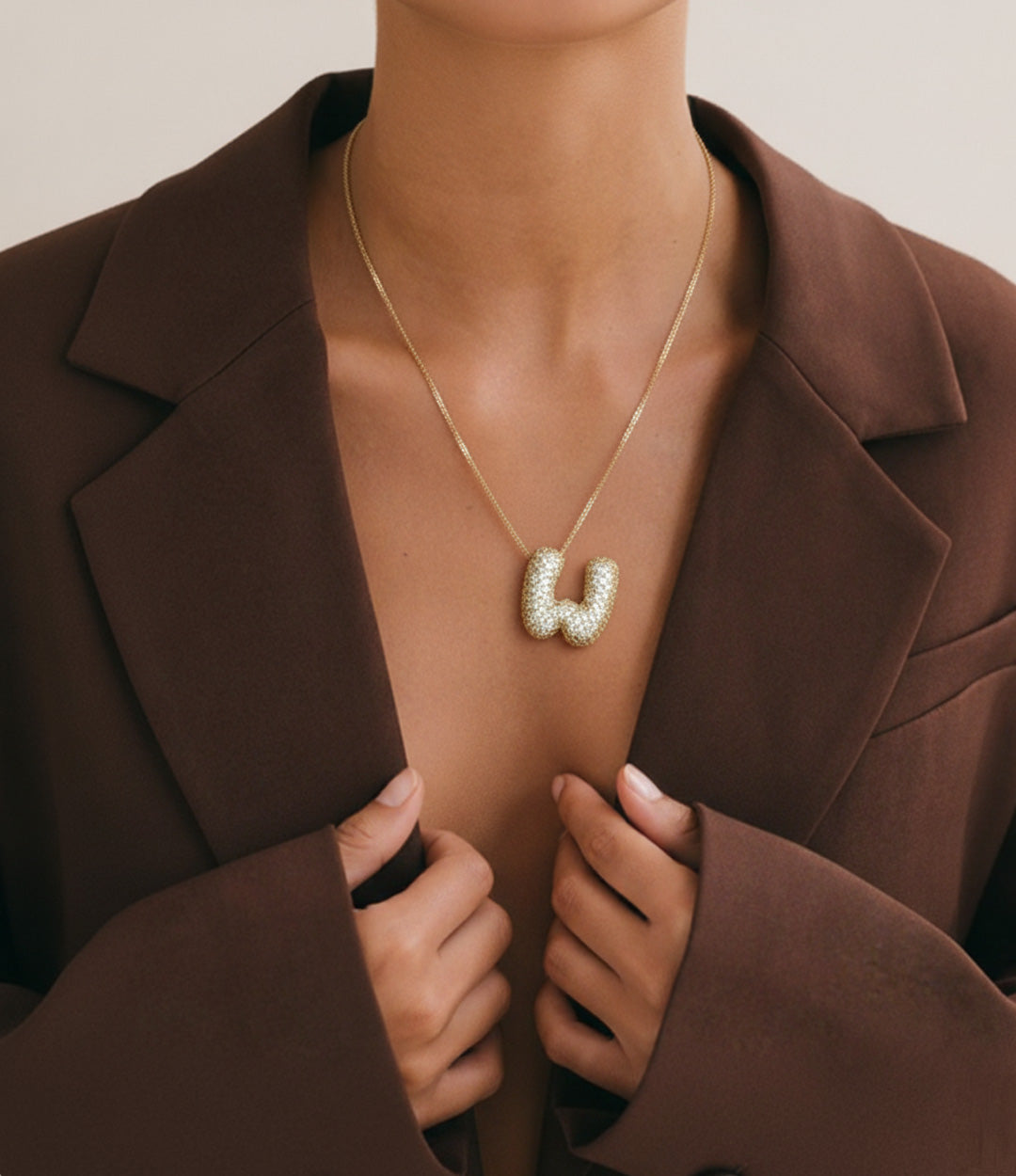 Close-up of a woman wearing a gold initial pendant necklace with a bubble letter design, styled with an open brown blazer.