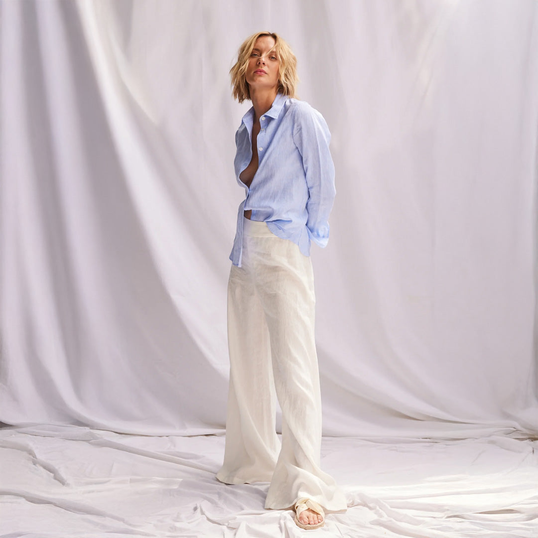 Woman wearing a light blue shirt and white pants against a white curtain background
