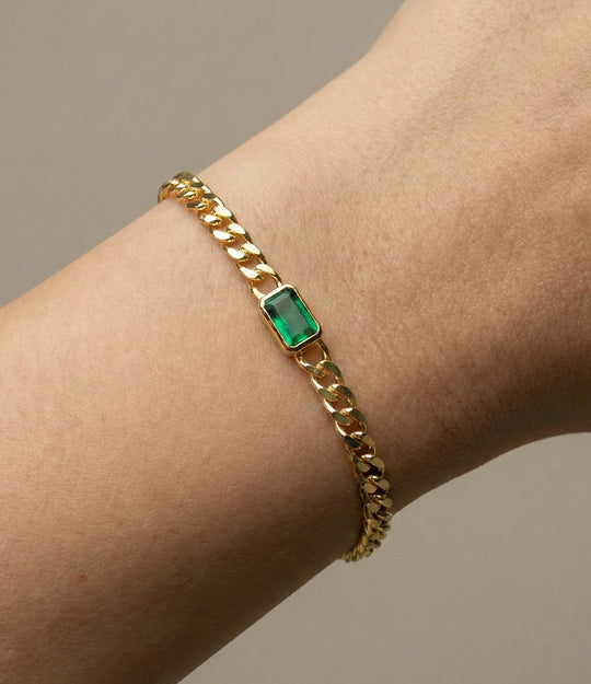 Gold chain bracelet with a green gemstone on a wrist against a neutral background
