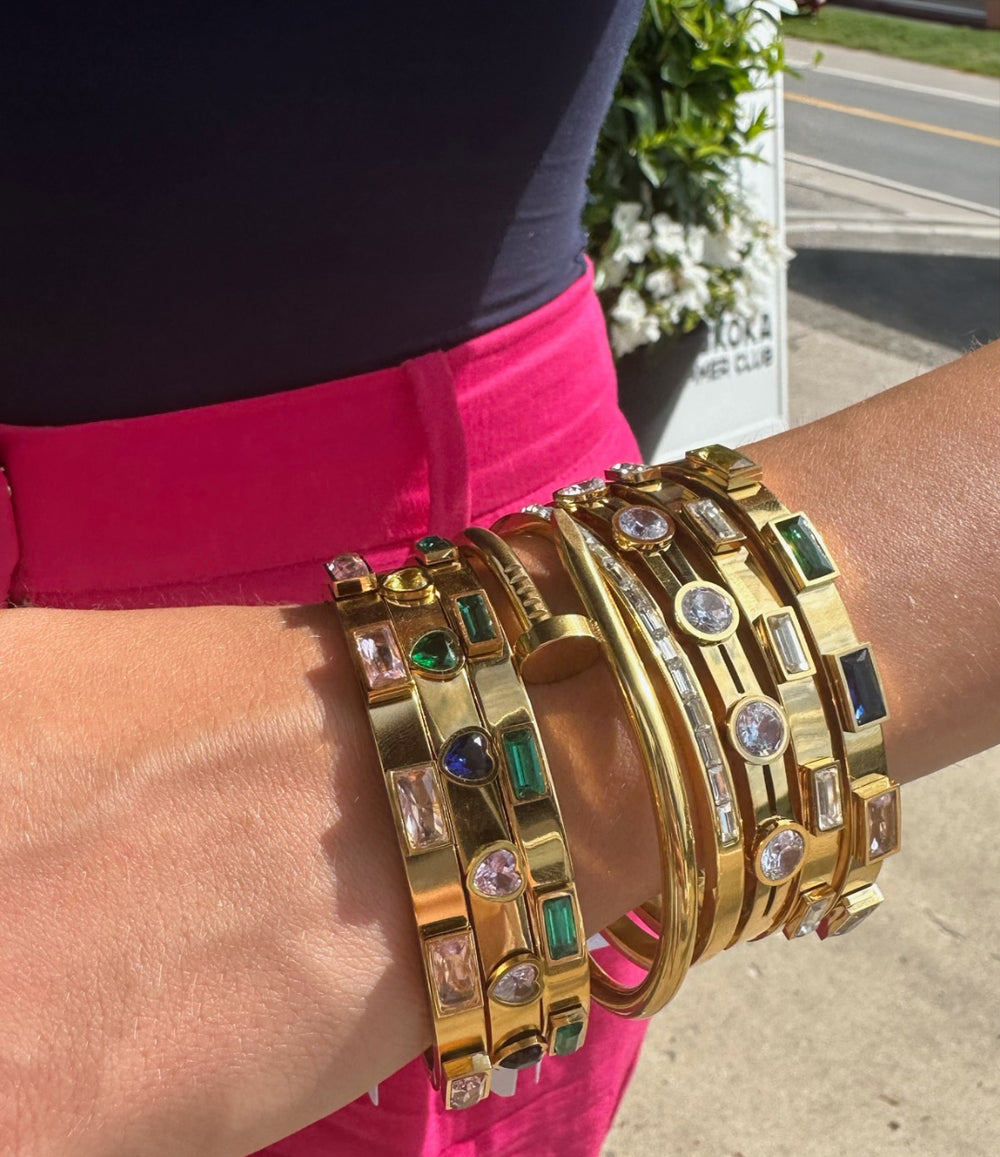 Gold bracelets with gemstones on a wrist, with a blurred background of a street and greenery.