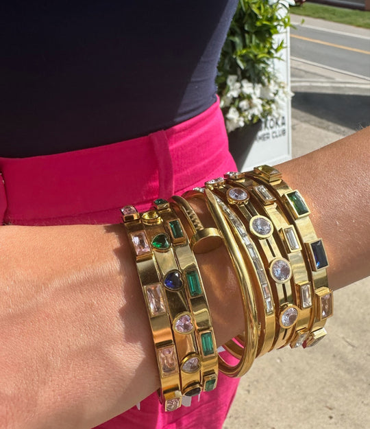Gold bracelets with gemstones on a wrist, with a blurred background of a street and greenery.