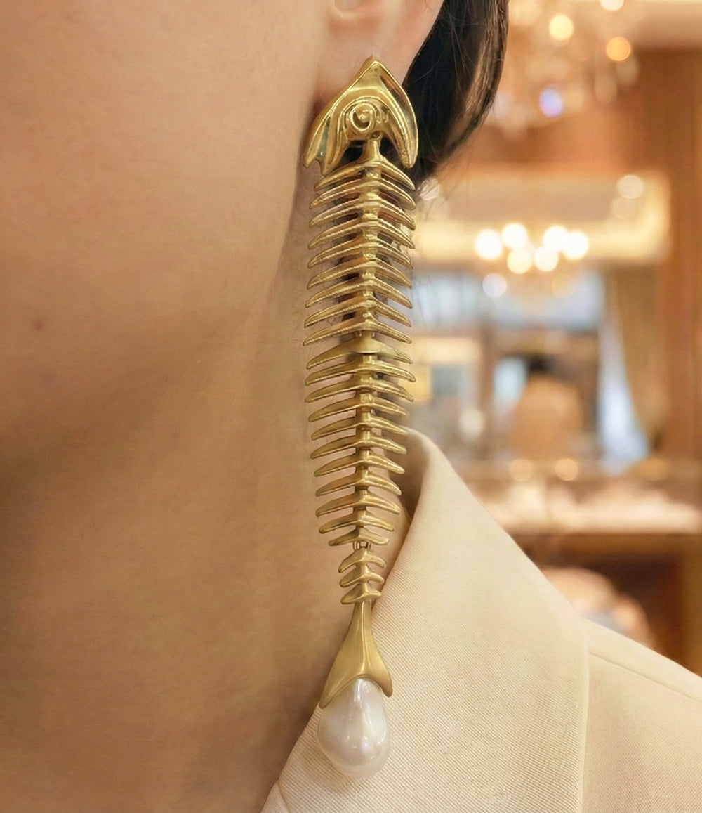 Gold earring with a pearl drop worn by a person, blurred indoor background