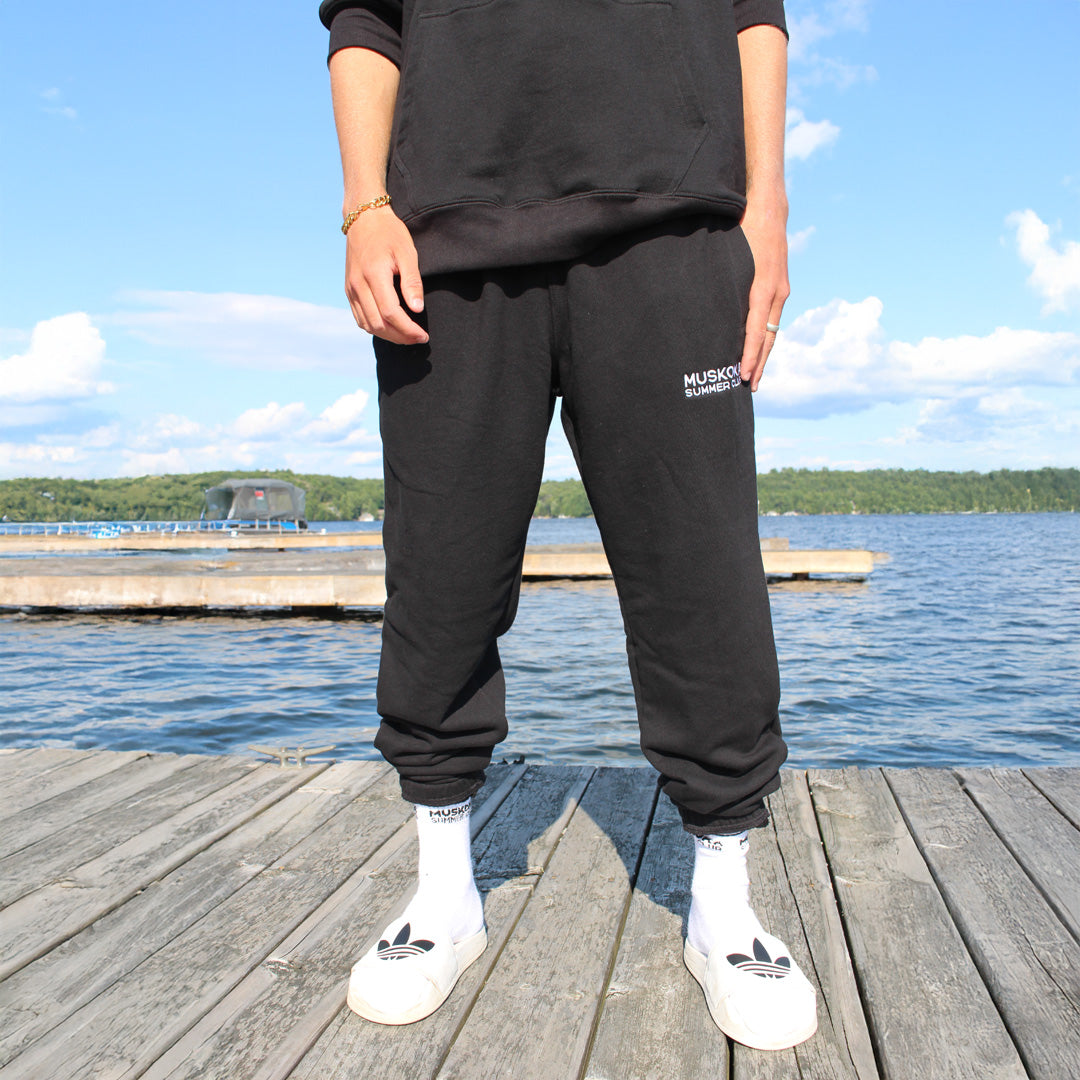 Person wearing black sweatpants with 'Muskoka Summer Club' branding and white sneakers on a dock by a lake.