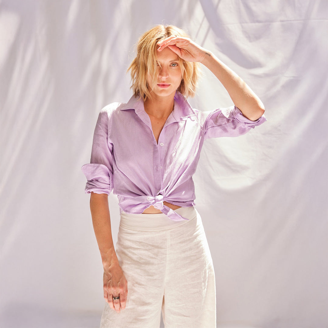 Woman wearing a light purple button-up shirt and white pants against a plain background