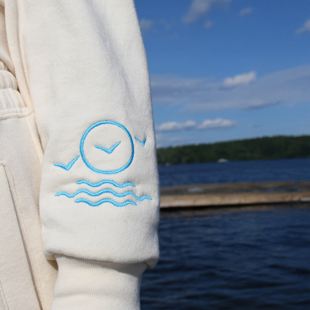 Light-colored sweatshirt with blue embroidery against a lakeside background