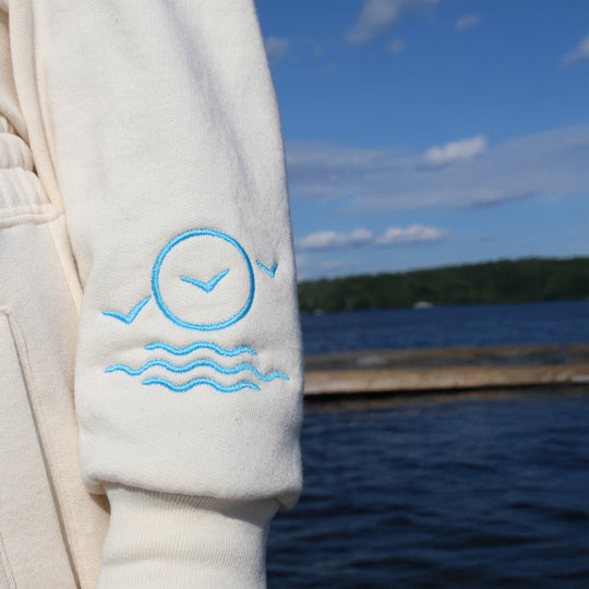 Light-colored sweatshirt with blue embroidery against a lakeside background