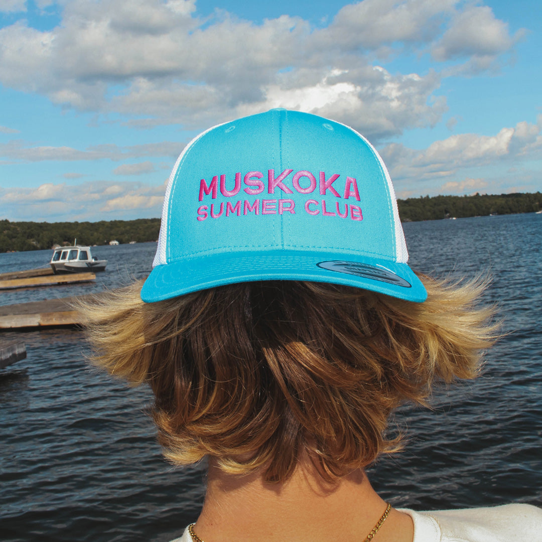 Person wearing a blue and white cap with 'Muskoka Summer Club' text, standing by a lake.